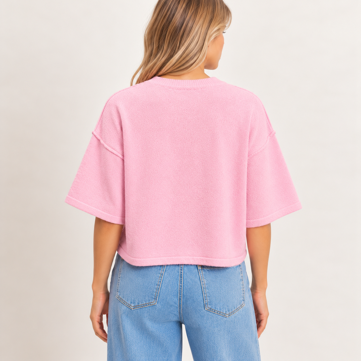 Back view of a pastel pink short sleeve knit sweater featuring a relaxed fit, dropped shoulders, ribbed crew neckline, and straight hem, styled with light-wash denim jeans.