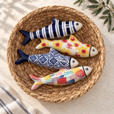 Four hand-painted stoneware fish in navy stripe, yellow floral, blue tile, and multicolor designs arranged in a woven basket