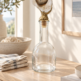 Clear glass bottle with a gold-tone oyster shell stopper displayed on a sunlit wood sideboard.