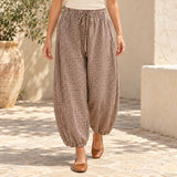 Model wearing brown gingham barrel-leg pants with an ivory top and tan flats in a coastal courtyard