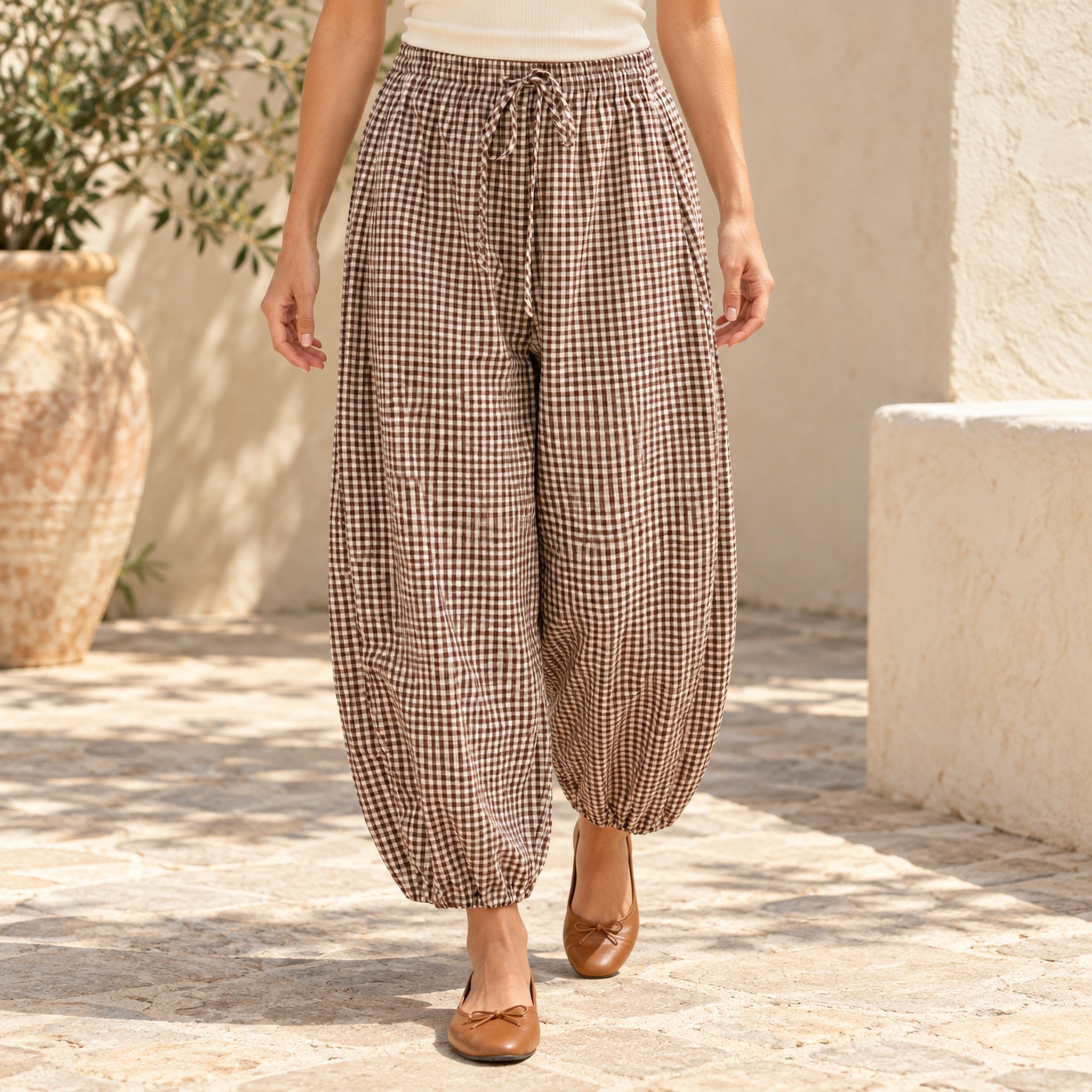 Model wearing brown gingham barrel-leg pants with an ivory top and tan flats in a coastal courtyard