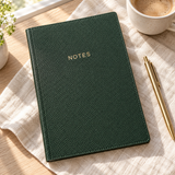 Forest green vegan leather notes journal on a linen-covered desk beside coffee and a gold pen.