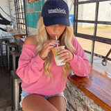 Woman wearing a 'unathletic' cap and pink sweatshirt, drinking iced coffee indoors.