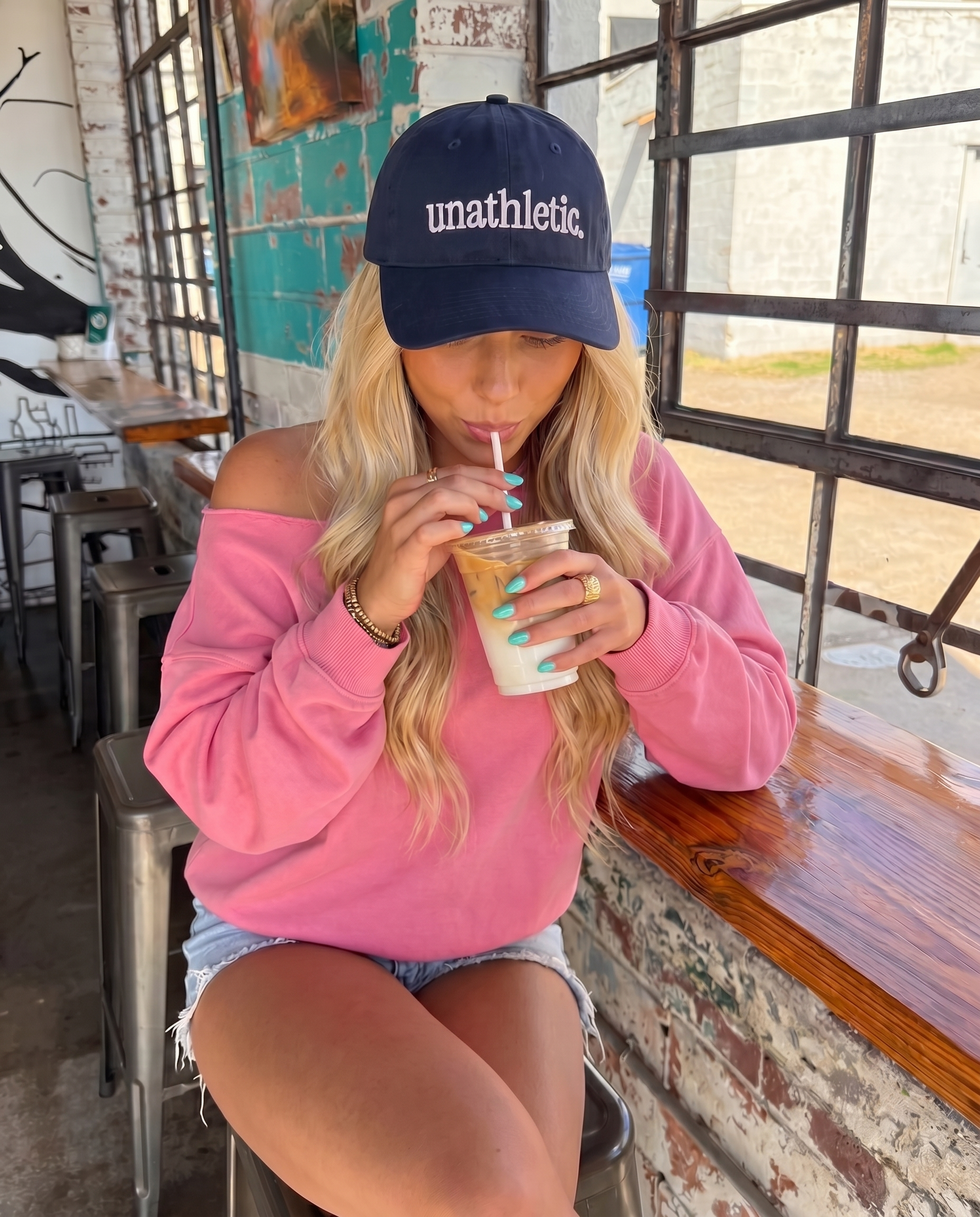 Woman wearing a 'unathletic' cap and pink sweatshirt, drinking iced coffee indoors.